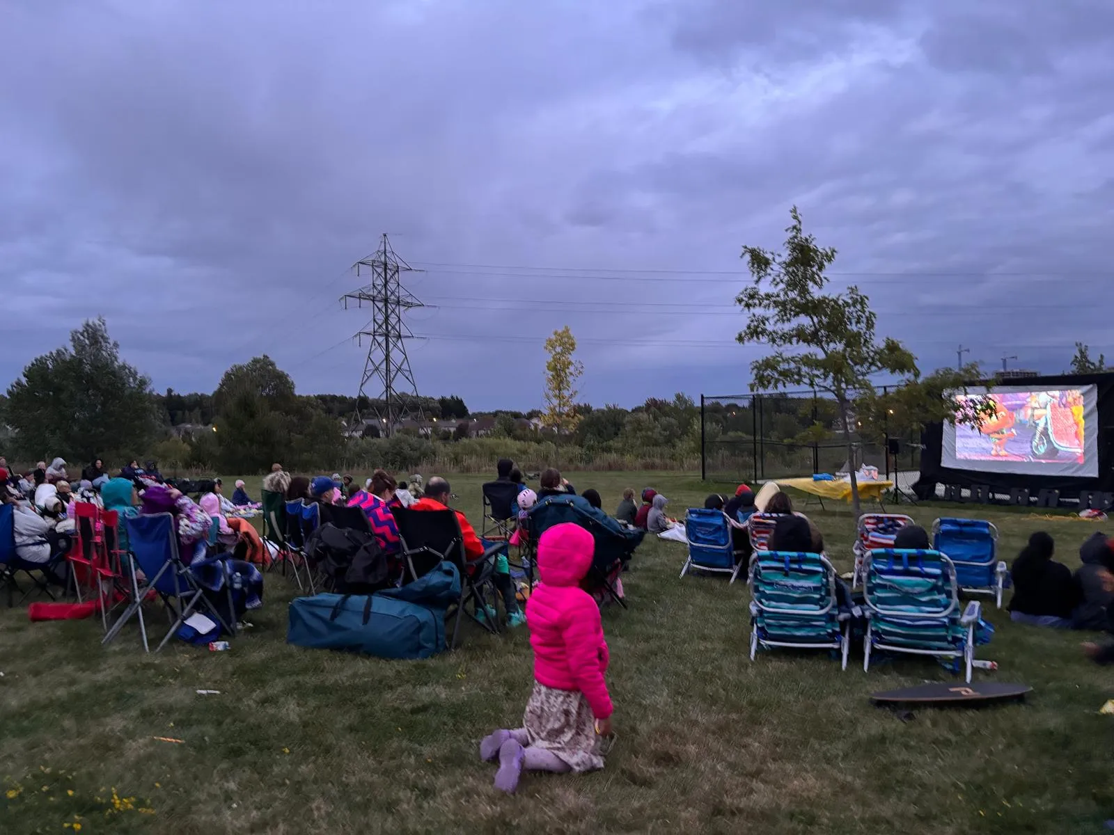 Neighbours in lawn chairs and on blankets watching a film on a big outdoor screen at dusk.