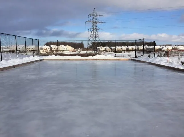 The volunteer-built outdoor rink at St. Moritz Park, boards up and ice in, with houses beyond on a snowy day.